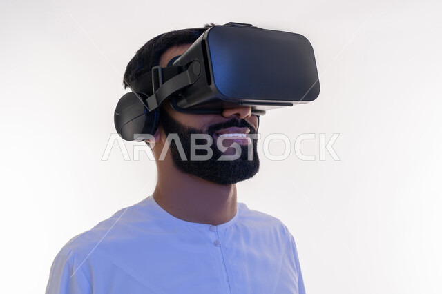 Technical and technological development, fun and entertainment watching through virtual reality glasses, close-up portrait of an Arab Gulf Emirati man wearing a kandura looking through 3D glasses, integration and simulation of the imaginary world, gestures of happiness and pleasure, white background