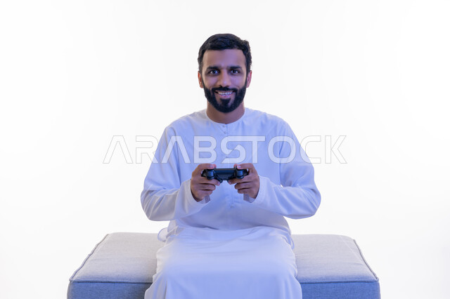 قضاء أوقات الفراغ بلعب ألعاب الفيديو الترفيهية، استخدام الأجهزة التكنولوجية والتقنيات الحديثة، بورتريه لرجل عربي خليجي اماراتي يرتدي الكندورة  الإماراتية يجلس على كرسي مريح يمسك بيده عصا التحكم، إيماءات الاستمتاع والاندماج والتفاعل, خلفية بيضاء