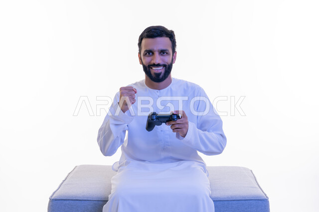رفع قبضة اليد بإيماءات الانتصار، استخدام الأجهزة التكنولوجية والتقنيات الحديثة، بورتريه لرجل عربي خليجي إماراتي مبتسم يرتدي الكندورة يجلس على كرسي مريح يمسك بيده عصا التحكم يقوم باللعب بألعاب الفيديو الترفيهية، خلفية بيضاء
