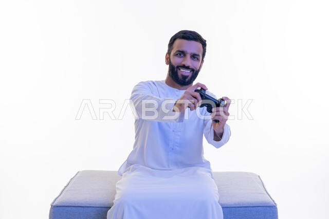 تعابير الاستمتاع والاندماج والتفاعل, قضاء أوقات الفراغ بلعب ألعاب الفيديو الترفيهية، استخدام الأجهزة التكنولوجية والتقنيات الحديثة، بورتريه لرجل عربي خليجي اماراتي يرتدي الكندورة  الإماراتية يجلس على كرسي مريح يمسك بيده عصا التحكم، خلفية بيضاء