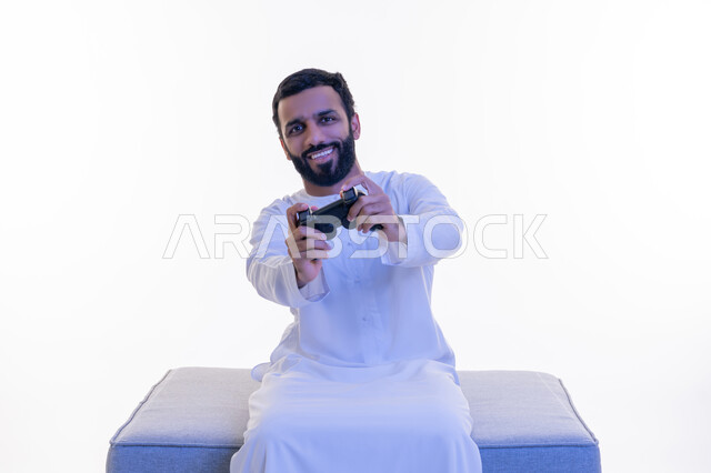 استخدام الأجهزة التكنولوجية والتقنيات الحديثة، بورتريه لرجل عربي خليجي اماراتي يرتدي الكندورة  الإماراتية يجلس على كرسي مريح يمسك بيده عصا التحكم، اللعب بألعاب الفيديو الترفيهية، إيماءات الاستمتاع والاندماج والتفاعل, خلفية بيضاء