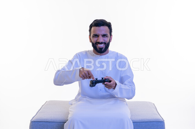 Gestures of enjoyment, integration and interaction, use of technological devices and modern techniques, portrait of an Arab Gulf Emirati man wearing a kandura sitting on a comfortable chair clicking on a joystick with expressions of enthusiasm, playing recreational video games, white background