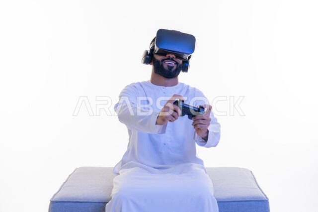 استخدام الأجهزة التكنولوجية والتقنيات الحديثة، بورتريه لرجل عربي خليجي إماراتي مبتسم يرتدي الكندورة ونظارة الواقع الإفتراضي يمسك بيده عصا التحكم يجلس على كرسي مريح يقوم باللعب بألعاب الفيديو الترفيهية، دمج العالم الخيالي مع المتعة والتسلية، خلفية بيضاء