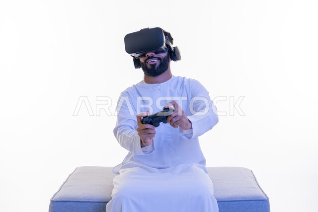 دمج العالم الخيالي مع المتعة والتسلية، استخدام الأجهزة التكنولوجية والتقنيات الحديثة، بورتريه لرجل عربي خليجي إماراتي مبتسم يرتدي الكندورة ونظارة الواقع الإفتراضي يمسك بيده عصا التحكم يجلس على كرسي مريح يقوم باللعب بألعاب الفيديو الترفيهية، خلفية بيضاء