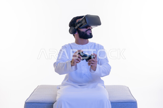 اللعب بألعاب الفيديو الترفيهية، بورتريه لرجل عربي خليجي إماراتي مبتسم يرتدي الكندورة ونظارة الواقع الإفتراضي يجلس على كرسي مريح يمسك بيده عصا التحكم، دمج العالم الخيالي مع المتعة والتسلية، استخدام الأجهزة التكنولوجية والتقنيات الحديثة، خلفية بيضاء