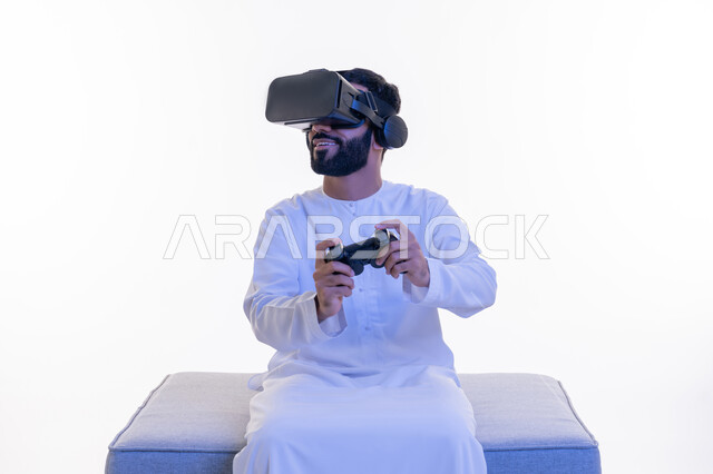 دمج العالم الخيالي مع المتعة والتسلية، الأجهزة التكنولوجية والتقنيات الحديثة، بورتريه لرجل عربي خليجي إماراتي مبتسم يرتدي الكندورة ونظارة الواقع الإفتراضي يمسك بيده عصا التحكم يجلس على كرسي مريح يقوم باللعب بألعاب الفيديو الترفيهية، خلفية بيضاء