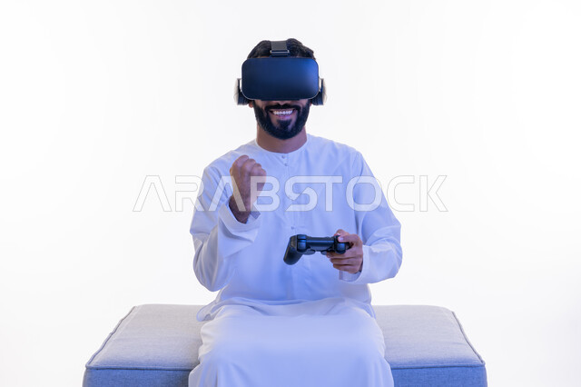 رفع قبضة اليد بإيماءات الحماس، دمج العالم الخيالي مع المتعة والتسلية، أجهزة تكنولوجية وتقنيات حديثة، بورتريه لرجل عربي خليجي إماراتي مبتسم يرتدي الكندورة ونظارة الواقع الإفتراضي يمسك بيده عصا التحكم يجلس على كرسي مريح يقوم باللعب بألعاب الفيديو، خلفية بيضاء