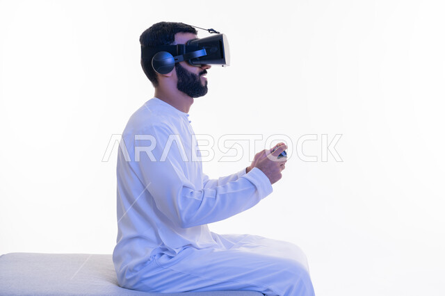 الأجهزة التكنولوجية والتقنيات الحديثة، بورتريه من الجانب لرجل عربي خليجي إماراتي يرتدي الكندورة ونظارة الواقع الإفتراضي يمسك بيده عصا التحكم يجلس على كرسي مريح يقوم باللعب بألعاب الفيديو الترفيهية، دمج العالم الخيالي مع المتعة والتسلية، خلفية بيضاء
