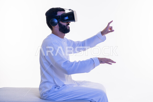 استخدام حاسة اللمس لملامسة عناصر هولوجرامية بإيماءات النقر, الاندماج والتفاعل ومحاكاة العالم الخيالي، بورتريه من الجانب لشاب عربي إماراتي يرتدي الكندورة ونظارة في آر الافتراضية ثلاثية الأبعاد يجلس على كرسي مريح يحاول الامساك بشيء ما، خلفية بيضاء