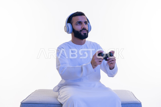 Using technological devices and modern techniques, portrait of an Arab Gulf Emirati man wearing a kandura, putting on headphones, sitting on a comfortable chair holding a joystick in his hand, playing entertaining video games, gestures of enjoyment, integration and interaction, white background