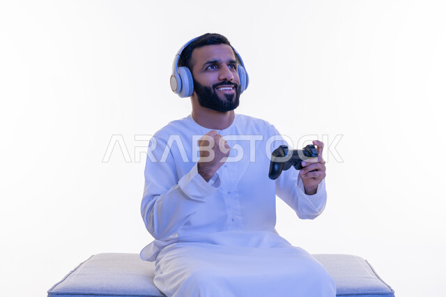 رفع قبضة اليد للأعلى بتعابير الانتصار والفوز، استخدام الأجهزة التكنولوجية والتقنيات الحديثة، بورتريه لرجل عربي خليجي اماراتي يرتدي الكندورة يضع سماعات الرأس يجلس على كرسي مريح يمسك بيده عصا التحكم، اللعب بألعاب الفيديو الترفيهية، خلفية بيضاء