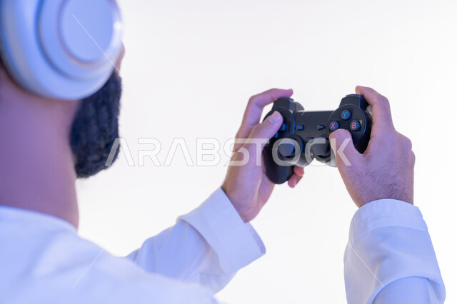 اللعب بألعاب الفيديو الترفيهية, استخدام الأجهزة التكنولوجية والتقنيات الحديثة، بورتريه مقرب من الجانب لرجل عربي خليجي اماراتي يرتدي الكندورة  الإماراتية يضع سماعات الرأس يمسك بيديه عصا التحكم، قضاء أوقات الفراغ بأنشطة ممتعة، خلفية بيضاء