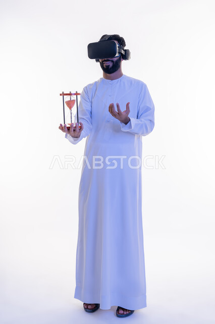 مفهوم إدارة الأعمال وتنظيم الجدول الزمني, إدراك قيمة اللحظات الثمينة, استخدام الأجهزة التكنولوجية والتقنيات الحديثة، بورتريه لرجل عربي خليجي اماراتي يرتدي الكندورة ونظارة في آر الافتراضية يمسك بيده الساعة الرملية الزجاجية، الجسم بالطول الكامل، خلفية بيضاء