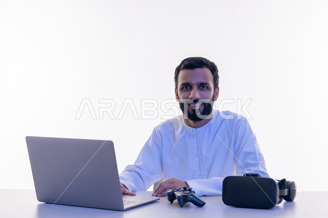 قضاء أوقات الفراغ بالقيام بأنشطة ترفيهية مسلية، استخدام الأجهزة التكنولوجية والتقنيات الحديثة، بورتريه مقرب لرجل عربي خليجي اماراتي يرتدي الكندورة يجلس خلف مكتبه أمامه نظارة الواقع الافتراضي وعصا التحكم يقوم بالعمل على الحاسوب المحمول، خلفية بيضاء