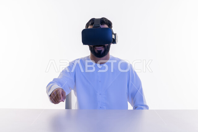 Expressions of happiness and pleasure, integration and interaction and simulation of the imaginary world, close-up portrait of a young Saudi Emirati Arab man wearing a kandura and 3D virtual VR glasses pointing at something with his index finger, using the sense of touch to touch holographic elements with tapping gestures, white background