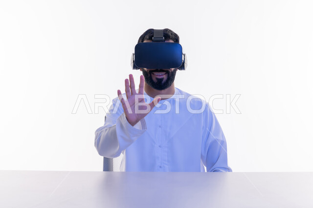 المتعة والتسلية بالمشاهدة عبر نظارة الواقع الافتراضي, بورتريه مقرب لرجل عربي خليجي إماراتي يرتدي الكندورة يضع نظارات ثلاثية الأبعاد يرفع يده في محاولة لمس شيء ما, إيماءات السعادة والسرور، الاندماج ومحاكاة العالم الخيالي، خلفية بيضاء