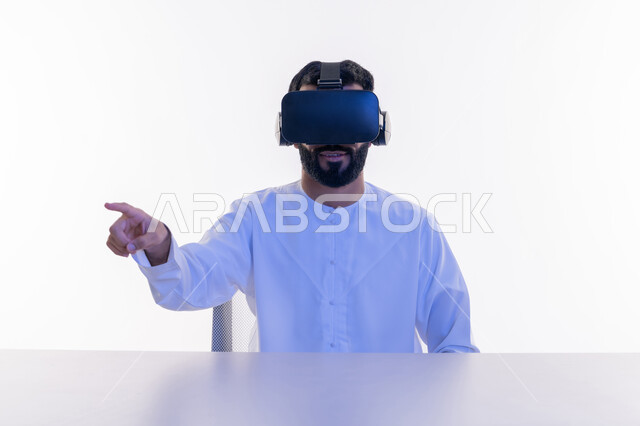 استخدام حاسة اللمس لملامسة عناصر هولوجرامية بإيماءات النقر, الاندماج والتفاعل ومحاكاة العالم الخيالي، بورتريه مقرب لشاب عربي سعودي اماراتي يرتدي الكندورة ونظارة في آر الافتراضية ثلاثية الأبعاد يشير إلى شيء ما بأصبعه السبابة، خلفية بيضاء