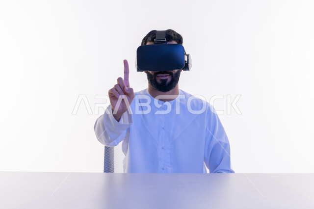 الاندماج والتفاعل ومحاكاة العالم الخيالي، بورتريه مقرب لشاب عربي سعودي اماراتي يرتدي الكندورة ونظارة في آر الافتراضية ثلاثية الأبعاد يشير إلى شيء ما بأصبعه السبابة، استخدام حاسة اللمس لملامسة عناصر هولوجرامية بإيماءات النقر, خلفية بيضاء