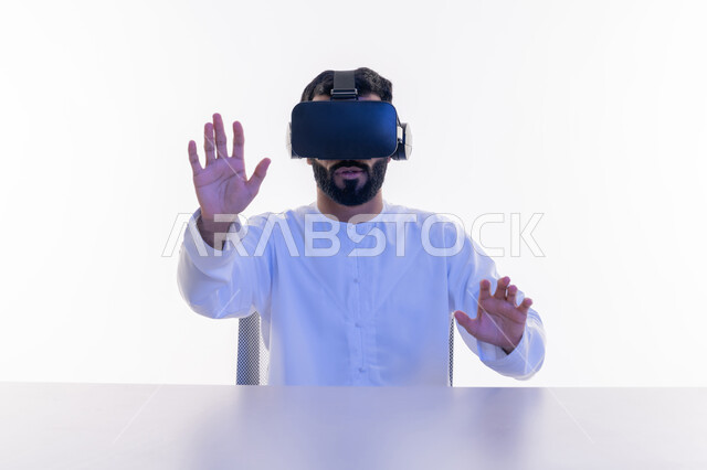 Gestures of happiness and pleasure, integration and simulation of the imaginary world, enjoyment and entertainment by watching through virtual reality glasses, close-up portrait of an Arab Gulf Emirati man wearing a kandura, putting on 3D glasses, raising his hands trying to touch something, white background