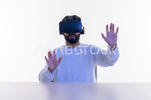 Integration and simulation of the imaginary world, fun and entertainment by watching through virtual reality glasses, close-up portrait of an Arab Gulf Emirati man wearing a kandura, putting on 3D glasses, raising his hands trying to touch something, gestures of happiness and pleasure, white background