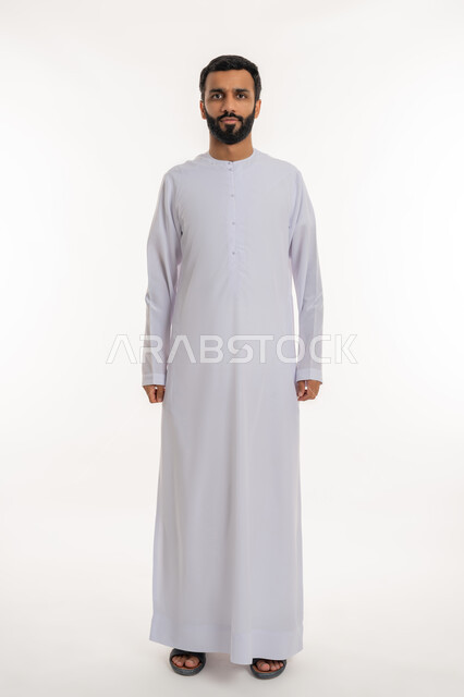 Looking at the camera with gestures of self-confidence, concept of elegance and masculinity, portrait of a smiling Emirati Gulf Arab man wearing a kandura standing straight with expressions of seriousness and firmness, interest in external appearance, full body length, white background