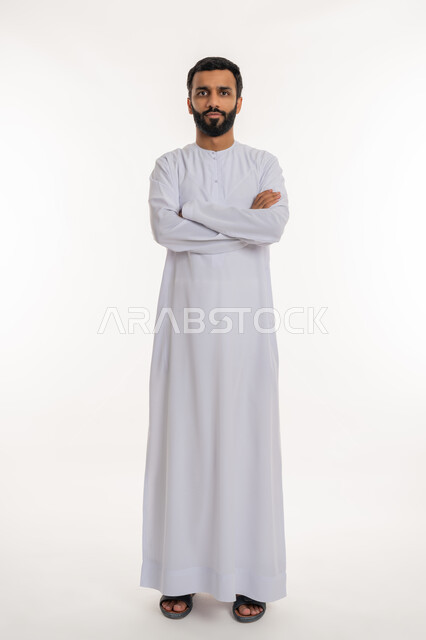 ايماءات الثقة بالنفس، مفهوم الاهتمام بالمظهر الخارجي و الرجولة والأناقه، بورتريه لرجل عربي خليجي إماراتي يرتدي الكندورة الإماراتية يقف باستقامة بايدي متقاطعه, النظر إلى الكاميرا بتعابيرالجدية والحزم، الطول الكامل للجسم، خلفية بيضاء