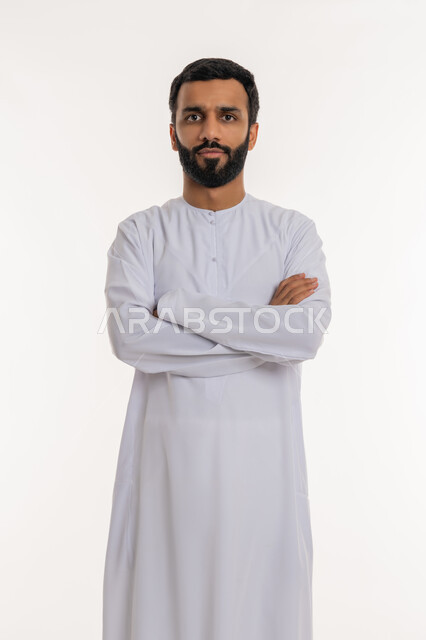 النظر إلى الكاميرا بتعابيرالجدية والحزم، ايماءات الثقة بالنفس، مفهوم الاهتمام بالمظهر الخارجي و الرجولة والأناقه، بورتريه لرجل عربي خليجي إماراتي يرتدي الكندورة الإماراتية يقف باستقامة بايدي متقاطعه, خلفية بيضاء