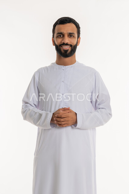 The concept of elegance and interest in appearance and masculinity, portrait of a smiling Emirati Gulf Arab man wearing a kandura standing with clasped hands, standing straight and looking at the camera with gestures of self-confidence and pleasure, white background