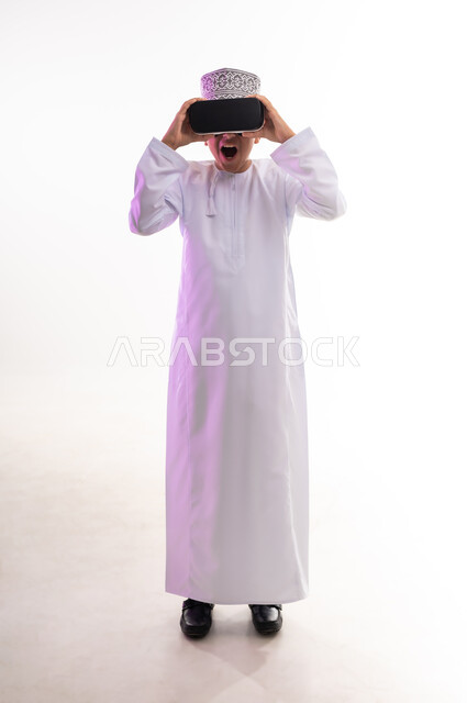 استخدام التقنيات والأجهزة المتطورة، محاكاة العالم الخيالي والنظر الى الافلام الواقعيه، المتعة والتسلية بالمشاهدة عبر نظارة الواقع الافتراضي،  بورتريه لولد عربي خليجي عماني يرتدي الدشداشه والكمه ينظر من خلال نظارات ثلاثية الأبعاد (الفي ار),  الجسم بالطول الكامل، خلفيه بيضاء