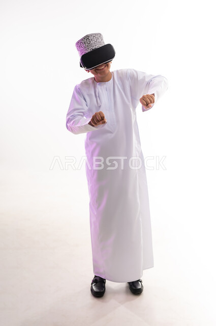 التسلية والمتعة بالمشاهدة عبر نظارة الواقع الافتراضي، استخدام التقنيات والأجهزة المتطورة، محاكاة العالم الخيالي والنظر الى الافلام الواقعيه، بورتريه لولد عربي خليجي عماني يرتدي الدشداشه والكمه ينظر من خلال نظارات ثلاثية الأبعاد (الفي ار),  الجسم بالطول الكامل، خلفيه بيضاء