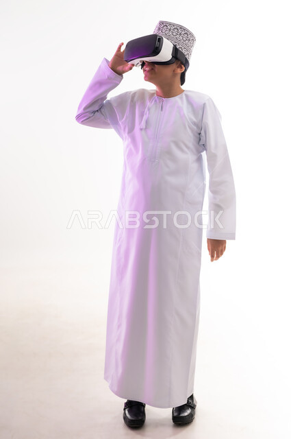 محاكاة العالم الخيالي والنظر الى الافلام الواقعيه، المتعة والتسلية بالمشاهدة عبر نظارة الواقع الافتراضي، التطور التقني والتكنولوجي,  بورتريه لولد عربي خليجي عماني يرتدي الدشداشه والكمه ينظر من خلال نظارات ثلاثية الأبعاد (الفي ار),  الجسم بالطول الكامل، خلفيه بيضاء