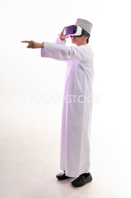استخدام التقنيات والأجهزة المتطورة، التسلية والمتعة بالمشاهدة عبر نظارة الواقع الافتراضي، بورتريه لولد عربي خليجي عماني يرتدي الدشداشه والكمه ينظر من خلال نظارات ثلاثية الأبعاد (الفي ار) ويشير باصبع السبابة الى مكان ما , الجسم بالطول الكامل، خلفيه بيضاء
