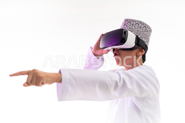 Using advanced technologies and devices, merging and simulating the imaginary world and looking at real movies, portrait of an Arab Gulf Omani boy wearing a dishdasha and a kilt looking through 3D glasses (VR) pointing with his index finger to a place, white background