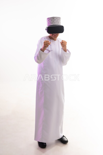 النظر الى الافلام الواقعيه ومحاكاة العالم الخيالي ، المتعة و التسلية بالمشاهدة عبر نظارة الواقع الافتراضي، التطور التقني والتكنولوجي, بورتريه لولد عربي خليجي عماني يرتدي الدشداشه والكمه ينظر من خلال نظارات ثلاثية الأبعاد (الفي ار)، الجسم بالطول الكامل، خلفيه بيضاء