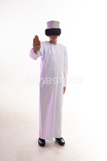 The concept of refusal and abstention, viewing through virtual reality glasses, portrait of an Arab Gulf Omani boy wearing a dishdasha and a cloak, looking through 3D glasses (VR), raising his palm up in gestures of forbidding doing something, requesting to stop and not approach, full-length body, white background