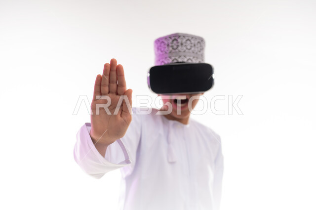 مفهوم الرفض والامتناع، المشاهدة عبر نظارة الواقع الافتراضي، طلب التوقف وعدم الاقتراب,  بورتريه لولد عربي خليجي عماني يرتدي الدشداشه والكمه ينظر من خلال نظارات ثلاثية الأبعاد (الفي ار) يرفع كفه للاعلى بإيماءات النهي عن فعل امر ما، خلفيه بيضاء