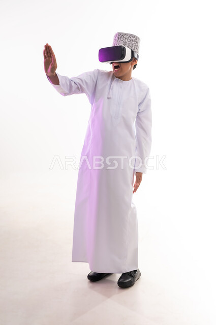 Viewing through virtual reality glasses, request to stop and not approach, concept of refusal and abstention, portrait of an Arab Gulf Omani boy wearing a dishdasha and a sleeve looking through 3D glasses (VR) raising his palm up in a gesture of forbidding doing something, full body, white background