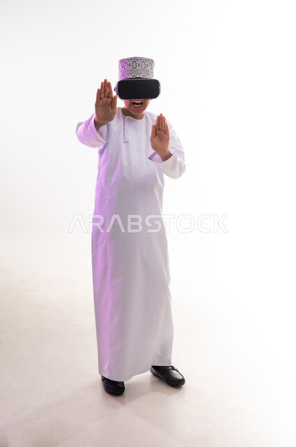 The concept of refusal and abstention, viewing through virtual reality glasses, portrait of an Arab Gulf Omani boy wearing a dishdasha and a cloak, looking through 3D glasses (VR), raising his palm up in gestures of forbidding doing something, requesting to stop and not approach, full-length body, white background