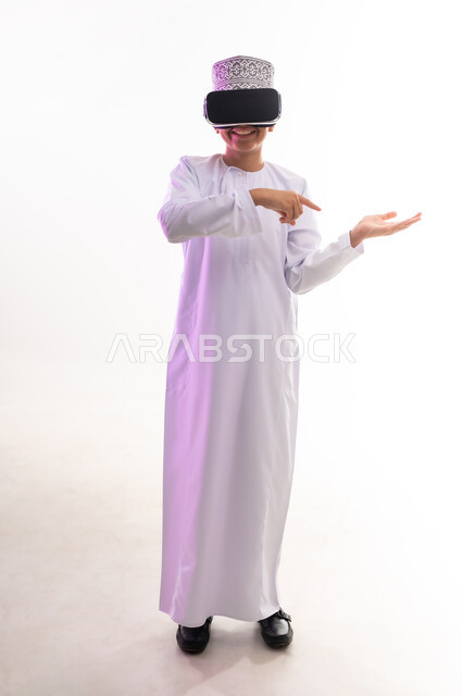 التسلية والمتعة بالمشاهدة عبر نظارة الواقع الافتراضي، استخدام التقنيات والأجهزة المتطورة، بورتريه لولد عربي خليجي عماني يرتدي الدشداشه والكمه ينظر من خلال نظارات ثلاثية الأبعاد (الفي ار) ويشير باصبع السبابة الى مكان ما ,  الجسم بالطول الكامل، خلفيه بيضاء