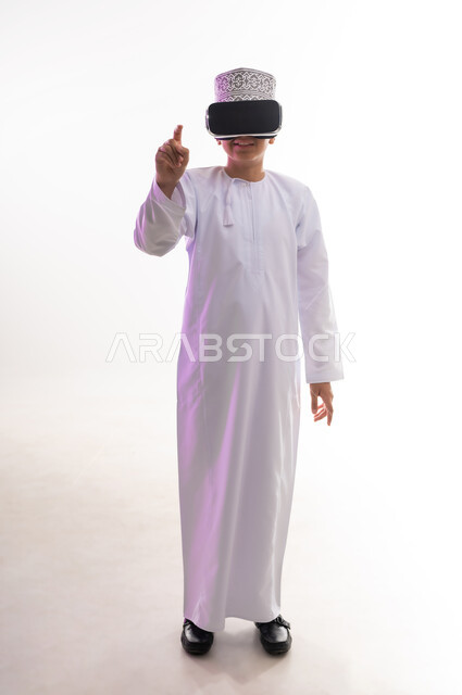 استخدام التقنيات والأجهزة المتطورة، التسلية والمتعة بالمشاهدة عبر نظارة الواقع الافتراضي، بورتريه لولد عربي خليجي عماني يرتدي الدشداشه والكمه ينظر من خلال نظارات ثلاثية الأبعاد (الفي ار) ويشير باصبع السبابة الى مكان ما , الجسم بالطول الكامل، خلفيه بيضاء