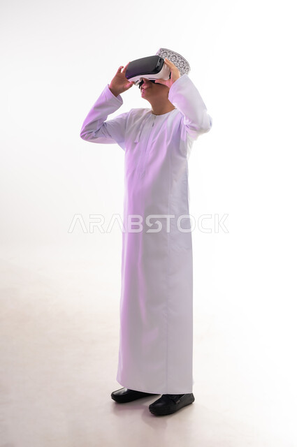 Integration and simulation of the imaginary world and watching real movies, enjoyment and entertainment by watching through virtual reality glasses, technical and technological development, portrait of an Arab Gulf Omani boy wearing a dishdasha and a cloak looking through 3D glasses (VR), full-length body, white background