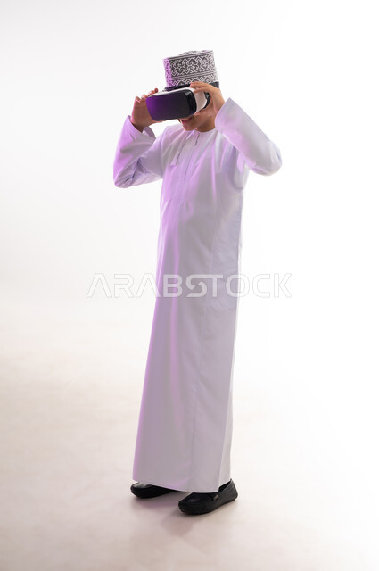 استخدام التقنيات والأجهزة المتطورة، محاكاة العالم الخيالي والنظر الى الافلام الواقعيه، المتعة والتسلية بالمشاهدة عبر نظارة الواقع الافتراضي،  بورتريه لولد عربي خليجي عماني يرتدي الدشداشه والكمه ينظر من خلال نظارات ثلاثية الأبعاد (الفي ار),  الجسم بالطول الكامل، خلفيه بيضاء
