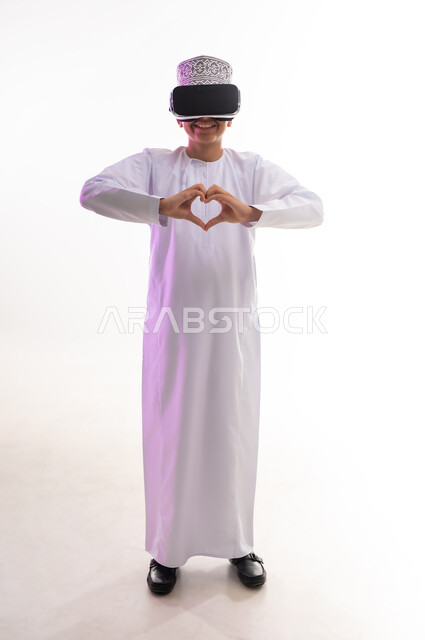 Expressions of love and affection, integration and simulation of the imaginary world, entertainment by watching through virtual reality glasses, technical and technological development, portrait of an Arab Gulf Omani boy wearing a dishdasha and a cloak looking through 3D glasses (VR) raising his hands in the shape of a heart, white background