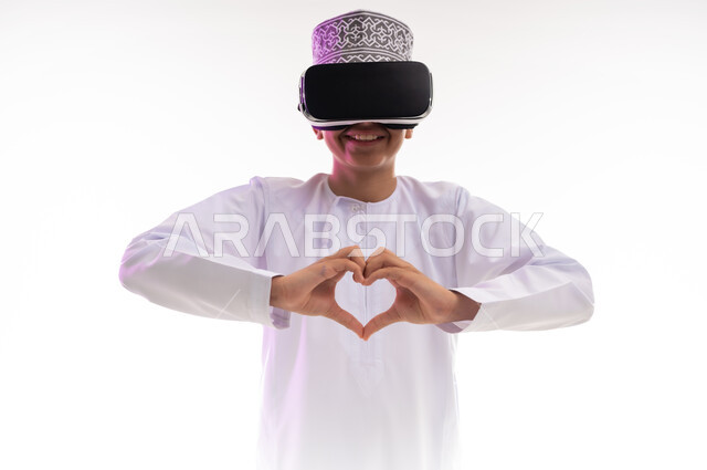 تعابير المحبة والمودة، الاندماج ومحاكاة العالم الخيالي، التسلية بالمشاهدة عبر نظارة الواقع الافتراضي، التطور التقني والتكنولوجي,  بورتريه لولد عربي خليجي عماني يرتدي الدشداشه والكمه ينظر من خلال نظارات ثلاثية الأبعاد (الفي ار) يرفع يديه على شكل قلب,  خلفيه بيضاء