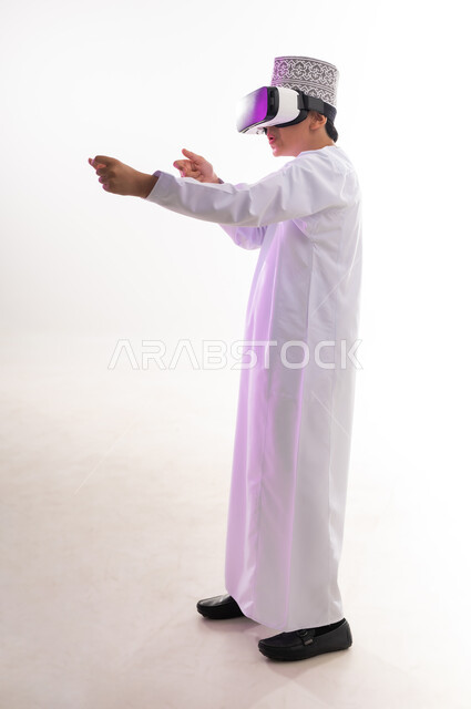 Raising hand with gestures of integration and simulation of the imaginary world, fun and entertainment by watching through virtual reality glasses, technical and technological development, portrait of an Arab Gulf Omani boy wearing a dishdasha and a cloak looking through 3D glasses (VR), full-length body, white background