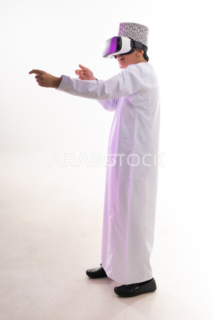 التطور التقني والتكنولوجي, رفع اليد بإيماءات الاندماج ومحاكاة العالم الخيالي، المتعة و التسلية بالمشاهدة عبر نظارة الواقع الافتراضي،  بورتريه لولد عربي خليجي عماني يرتدي الدشداشه والكمه ينظر من خلال نظارات ثلاثية الأبعاد (الفي ار),  الجسم بالطول الكامل، خلفيه بيضاء