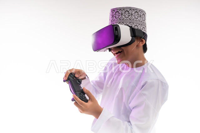 المتعة والتسلية وقضاء أوقات ممتعة في ألعاب الفيديو الترفيهيه، الاندماج ومحاكاة العالم الخيالي، استخدام الأجهزة والتقنيات الحديثة، بورتريه لولد عربي خليجي عماني يرتدي الدشداشه والكمه ينظر من خلال نظارات ثلاثية الأبعاد (الفي ار) يمسك بيده عصا التحكم, خلفيه بيضاء