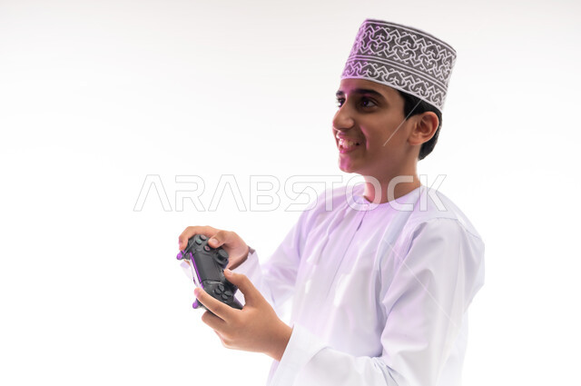قضاء أوقات ممتعة في ألعاب الفيديو الترفيهيه، بورتريه لولد عربي خليجي عماني يرتدي الدشداشة والكمة يحمل بيده عصا التحكم، إيماءات الاستمتاع والاندماج، استخدام الأجهزة التكنولوجية والتقنيات الحديثة، خلفية بيضاء
