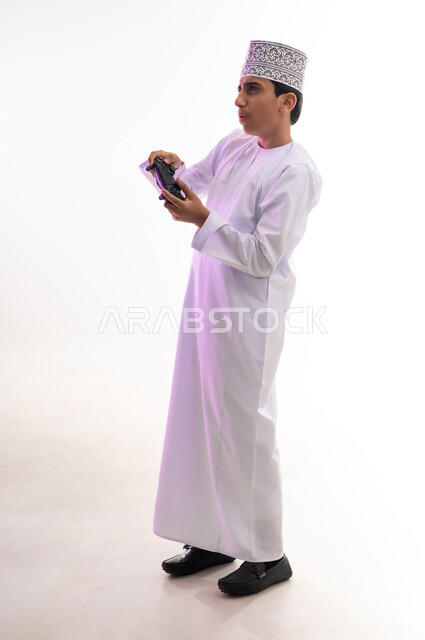 Having fun playing video games, portrait of an Omani Gulf Arab boy wearing a dishdasha and a kimono holding a joystick, gestures of enjoyment and integration, using technological devices and modern techniques, white background