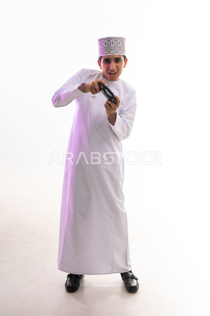 قضاء أوقات ممتعة في ألعاب الفيديو الترفيهيه، بورتريه لولد عربي خليجي عماني يرتدي الدشداشة والكمة يحمل بيده عصا التحكم، إيماءات الاستمتاع والاندماج، استخدام الأجهزة التكنولوجية والتقنيات الحديثة، خلفية بيضاء